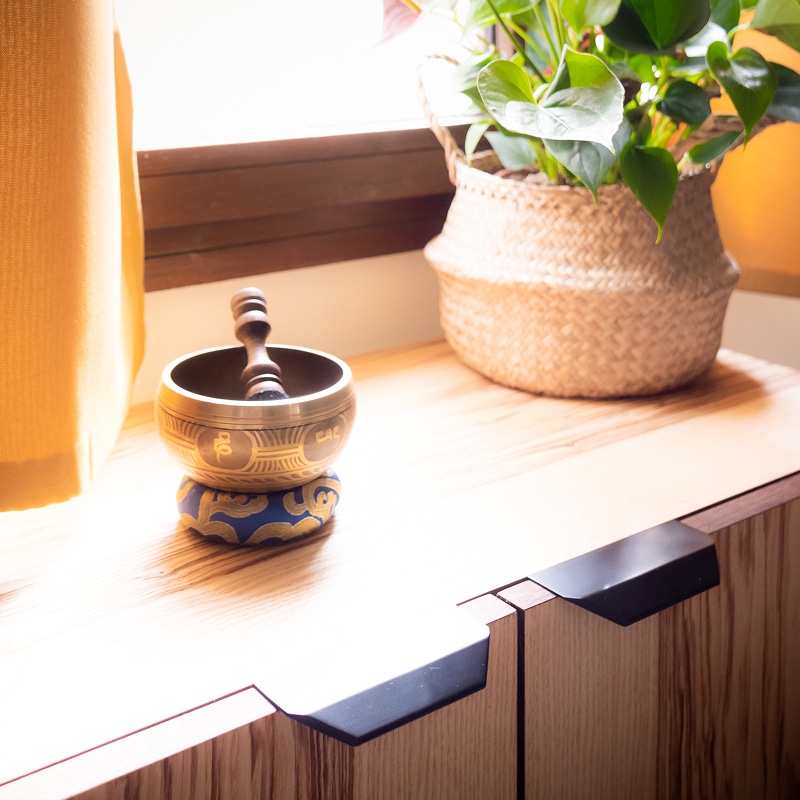 Bol chantant et ambiance zen sur meuble en bois Bol chantant tibétain en laiton sur un coussin bleu, posé sur un meuble en bois clair près d'une fenêtre avec un rideau jaune et une plante en panier.