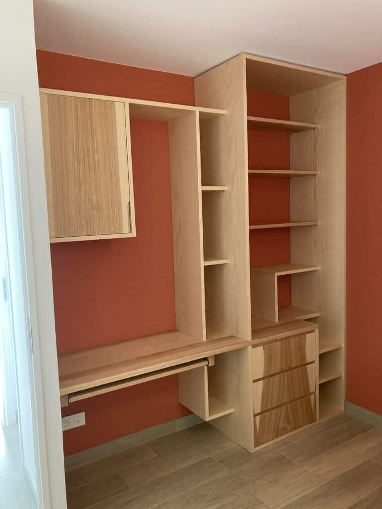 Bureau intégré en bois clair sur mur couleur terre cuite, avec placard suspendu, étagères et tiroirs.