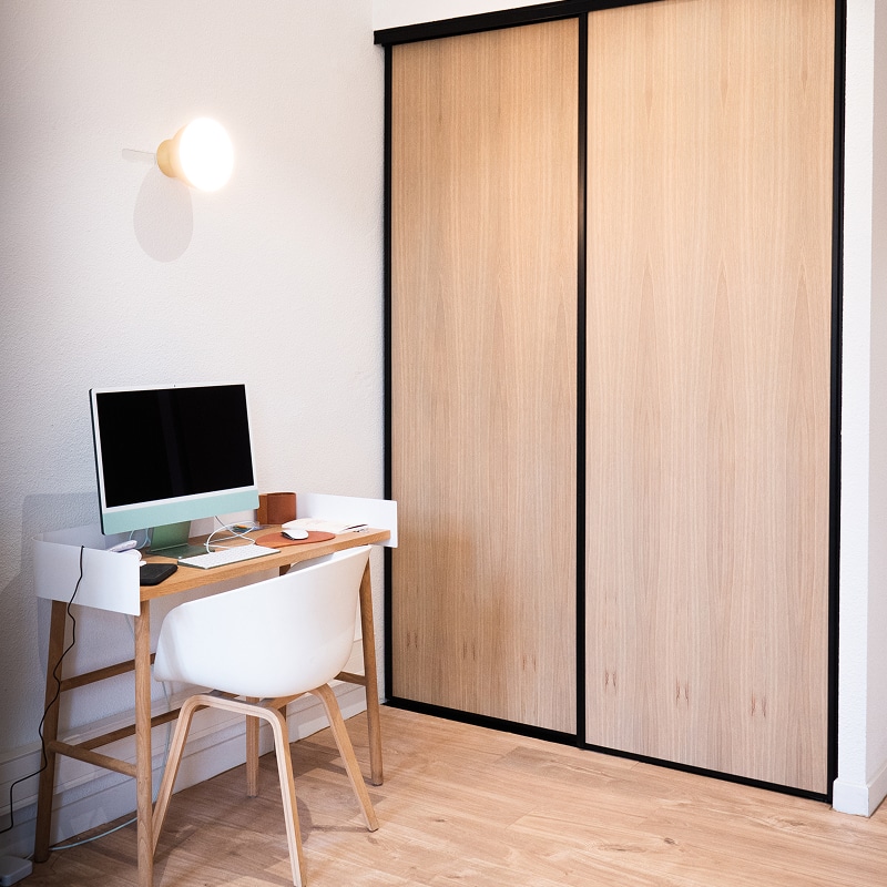 Bureau Design avec Rangement Coulissant Clair Coin bureau moderne avec un ordinateur iMac, une chaise blanche et un bureau en bois clair, à côté d'un grand placard à portes coulissantes en bois clair à cadre noir.