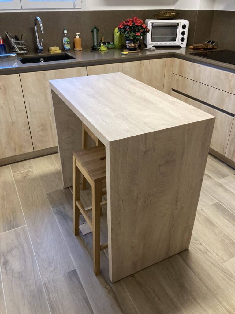 Îlot de cuisine moderne en bois clair avec tabouret, plan de travail sombre, évier et petit four électrique blanc en arrière-plan.