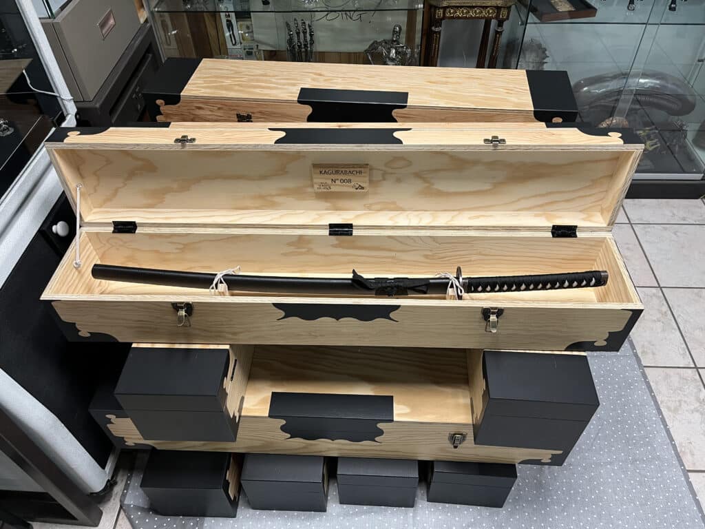 Katana Kagurabachi N° 008 : Pièce de Collection Ultime Katana Kagurabachi N° 008, édition limitée, exposé dans un coffret en bois clair et noir. Texte : "POUR SOMMES DES HÉROS".