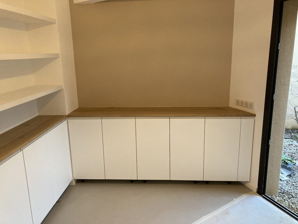 Rangement Cuisine Moderne : Meuble Bas et Comptoir Bois Meuble bas blanc moderne avec comptoir bois clair, étagères ouvertes et multiprise murale, près d'une porte-fenêtre extérieure.