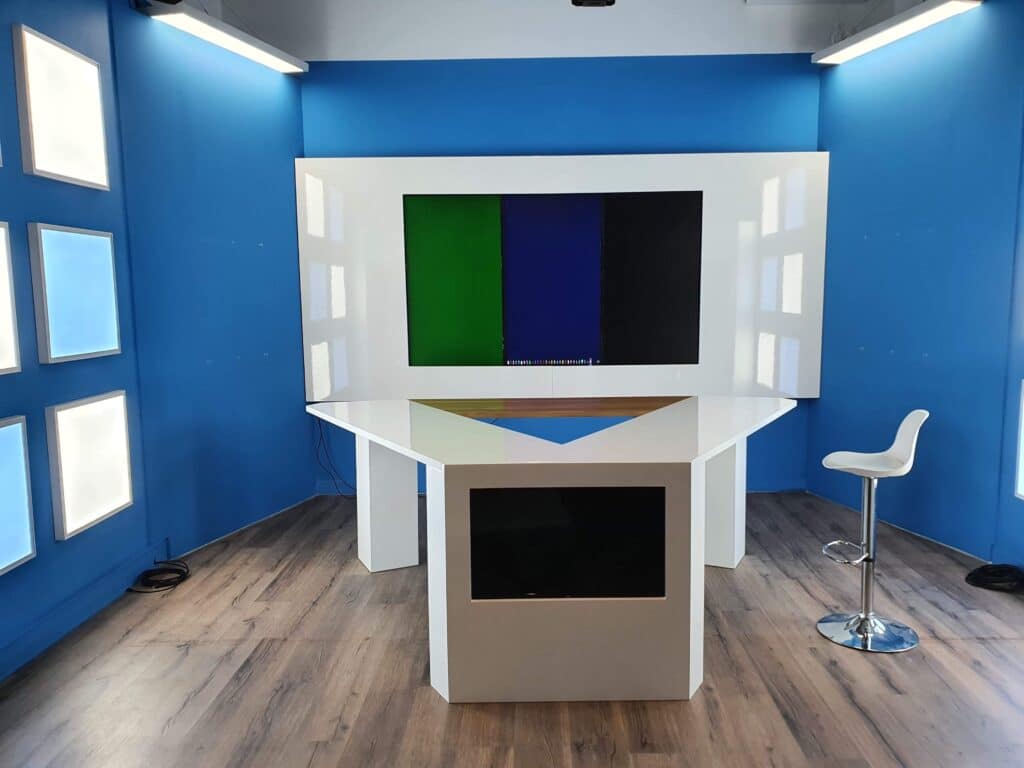 Studio moderne aux murs bleus avec des panneaux lumineux, un bureau blanc angulaire affichant un motif de barres de couleur (vert, bleu, noir) et un écran intégré.